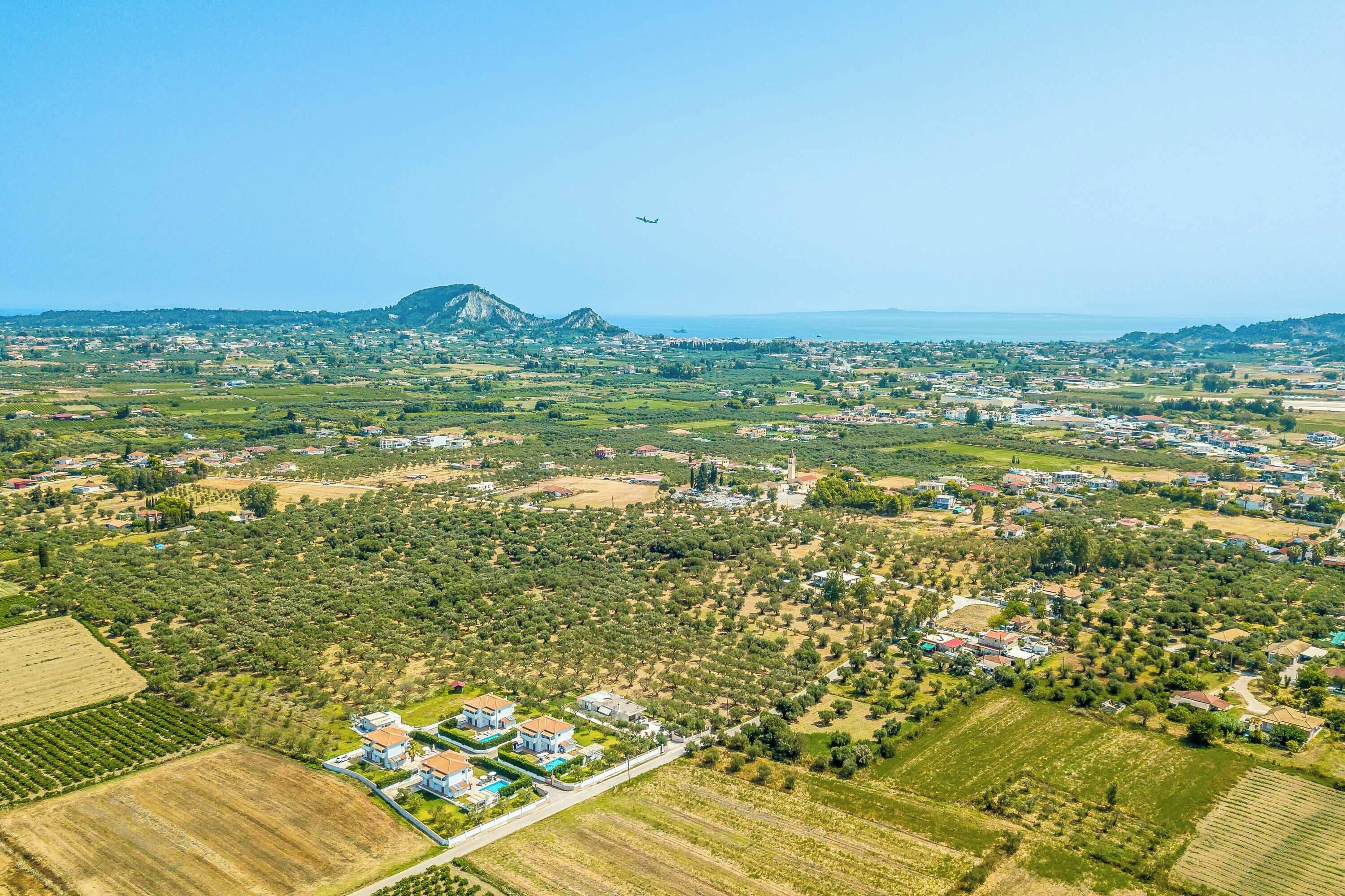 An aerial view of a coastal town with residential areas, olive groves, and a prominent mountain in the background.