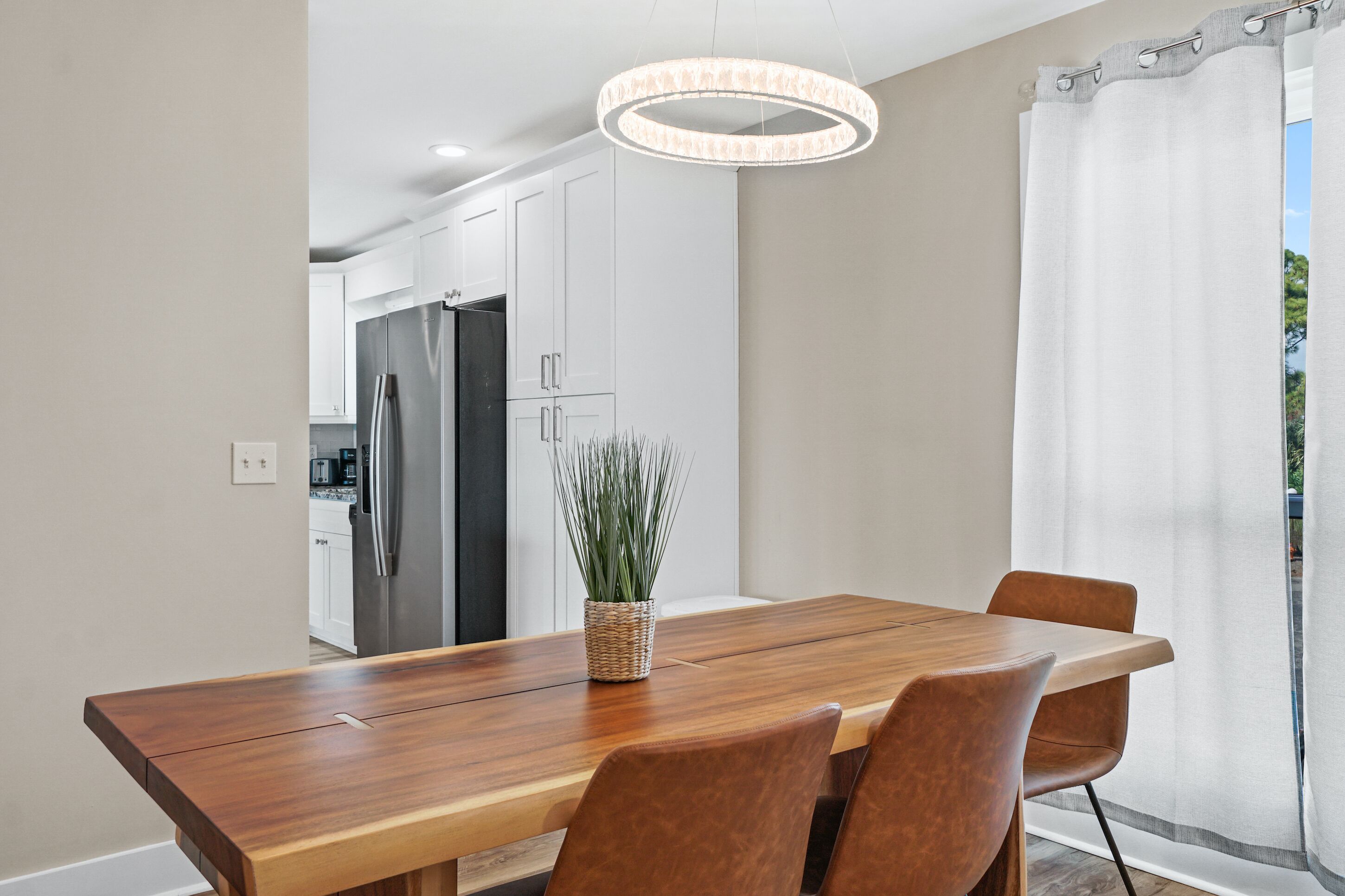Una cocina moderna con armarios blancos y electrodomésticos de acero inoxidable, una mesa de comedor de madera con sillas, y una lámpara colgante.