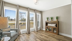 Ein geräumiges Wohnzimmer mit großen Fenstern, Holzboden, einer Couch, einer Lampe und einem Holzschrank mit Dekoration.