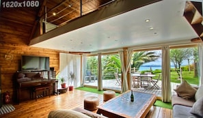 A spacious living area with a wooden floor, a sofa, a coffee table, and a TV. Large windows offer a view of the outdoors.