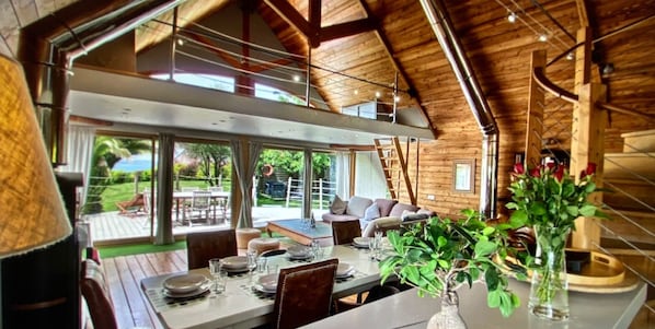 A spacious dining area with a high ceiling and wooden beams, overlooking a lush garden.