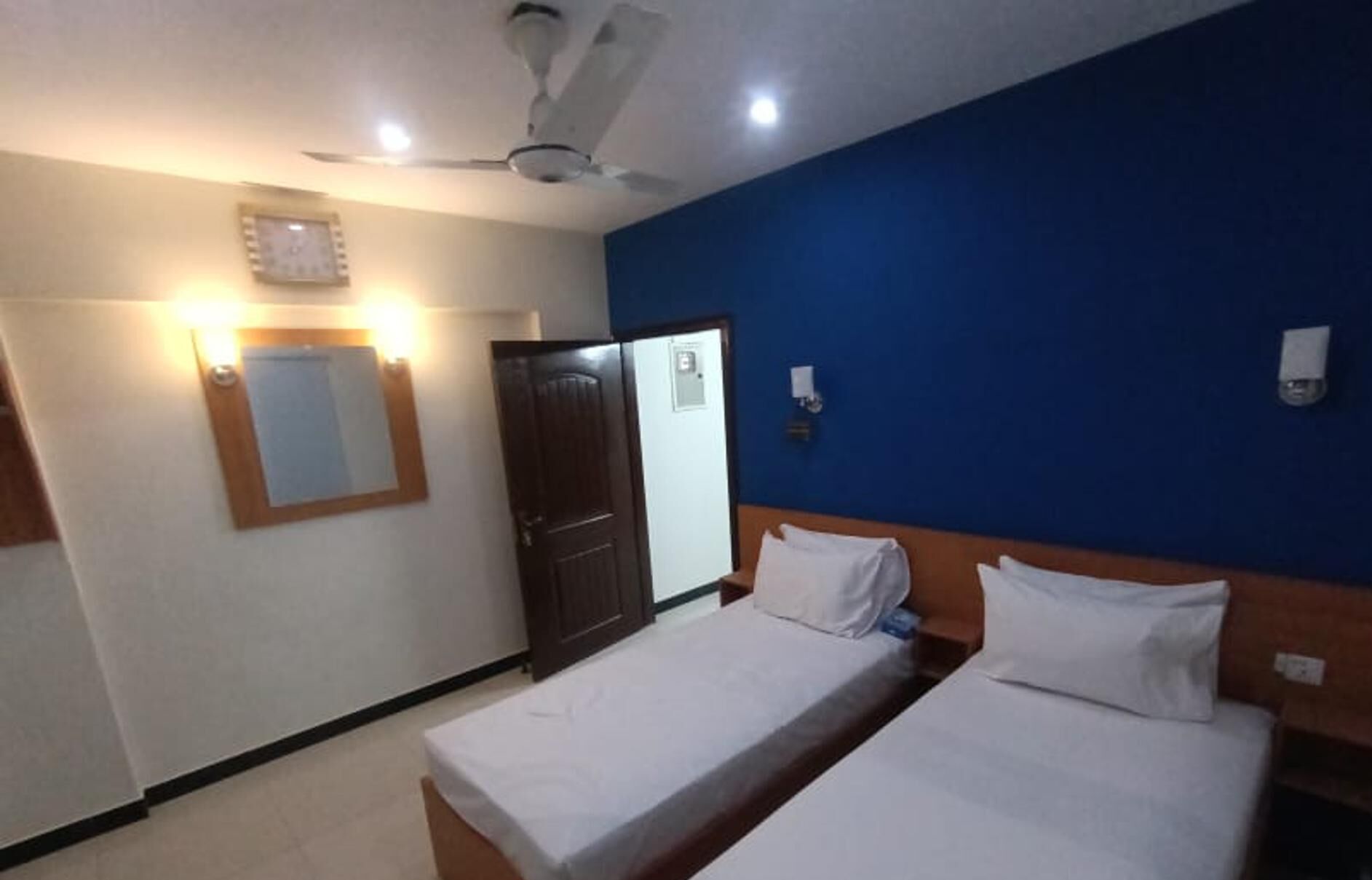 A hotel room with two single beds, a ceiling fan, a door, and a wall-mounted light fixture.