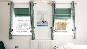 Una habitación con dos ventanas, cada una con una barra para cortinas y cortinas a juego. Se ve un radiador debajo de las ventanas, y un cuadro cuelga en la pared entre ellas.
