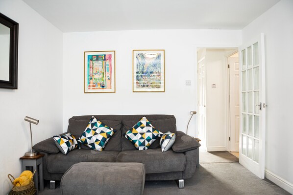 A living room with a grey sofa, patterned cushions, a side table, and a lamp. There are two framed artworks on the wall and a door leading to another room.