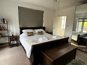 A bedroom with a large bed, a bedside table with a lamp, a bookshelf, and a mirror.