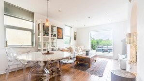Amplia sala de estar con una mesa redonda de vidrio, sillas traslúcidas, una mesa de centro de madera y vistas al exterior a través de grandes ventanales.