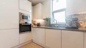 Una cocina moderna con armarios blancos, un horno de acero inoxidable, un microondas, una cafetera y una ventana que proporciona luz natural.