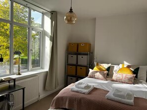 A bedroom with a large window, a bed with pink bedding, a bedside table, and storage boxes.