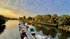 A calm river with boats docked along the pier, surrounded by trees and buildings.