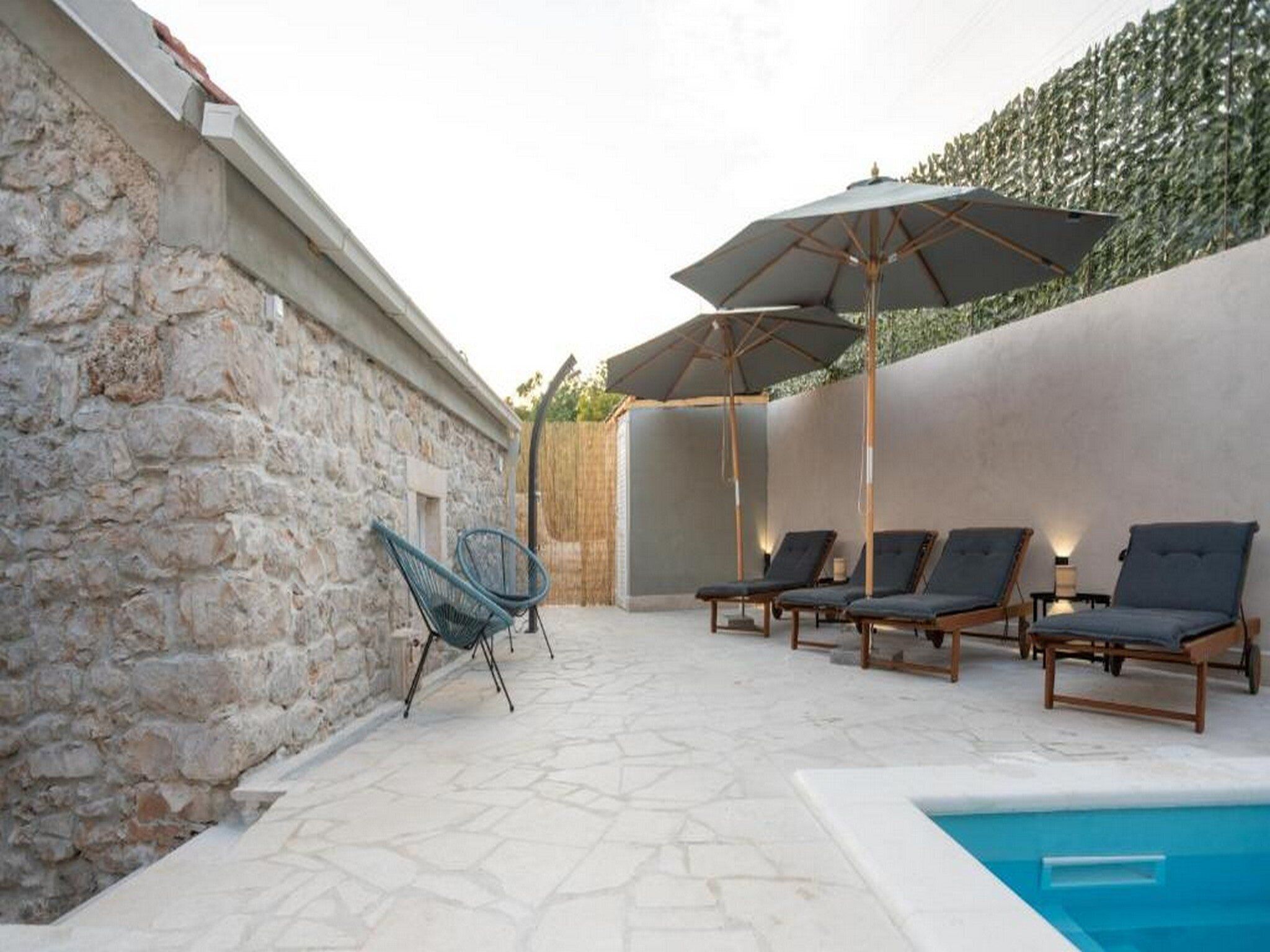 Zona de piscina con sillones de descanso y sombrillas, muros de piedra y una vista despejada a la piscina.