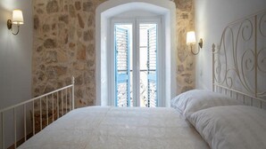 A bedroom with a stone wall, a white door with shutters, a bed with white linens, and wall-mounted light fixtures. - House of Nice Memories (Orebic)