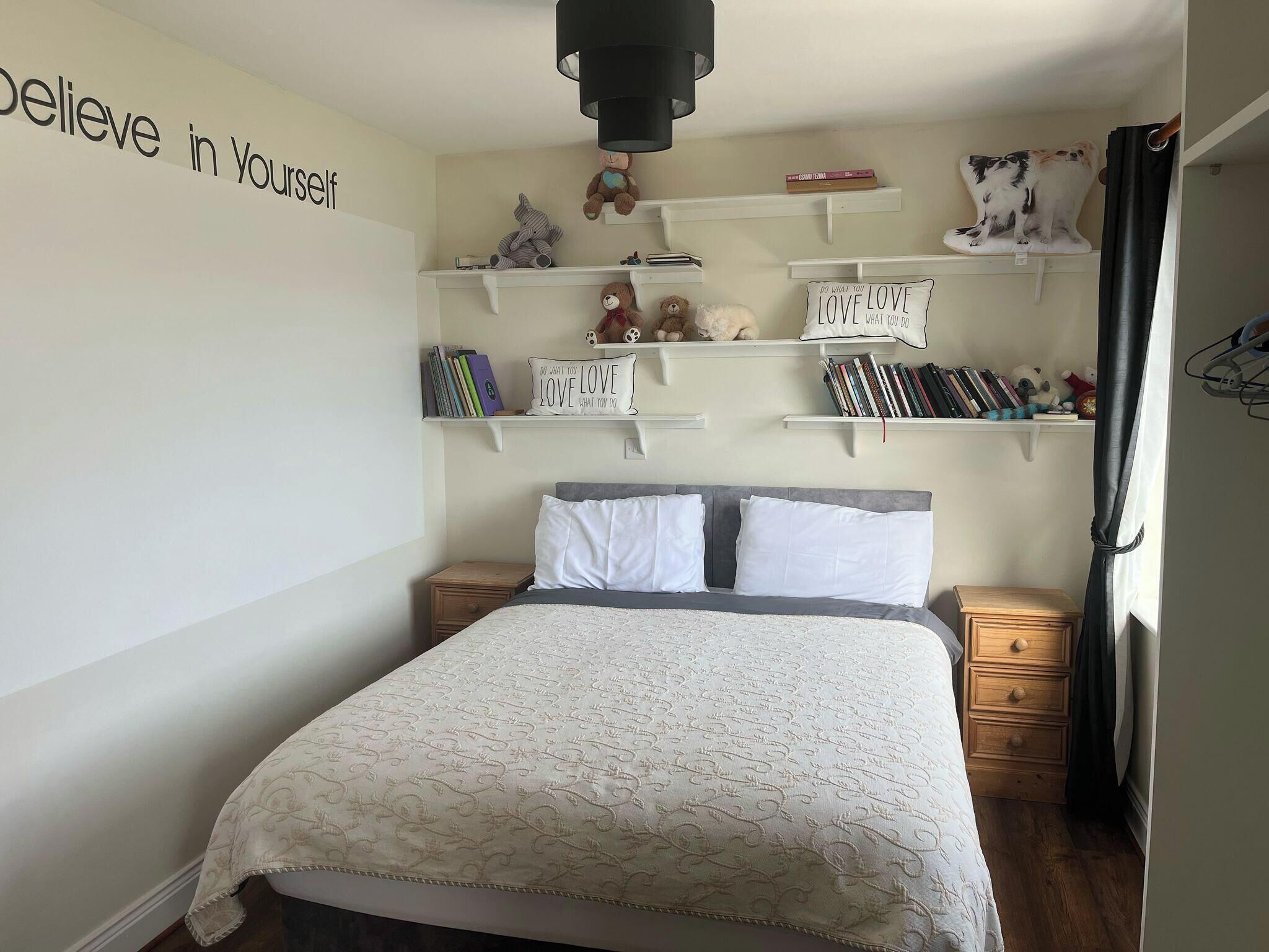 Ein Schlafzimmer mit einem Bett, Nachttischen, Regalen mit Büchern und Dekoration, einem motivierenden Wandspruch und einem Fenster mit Vorhängen.