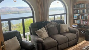 A living room with a sofa, two armchairs, a bookshelf, and large windows with a view of a lake and mountains. - 6 Ensuite Bedrooms-sleeps 12-garden-sea & Mtn View (Blackwater Bridge)