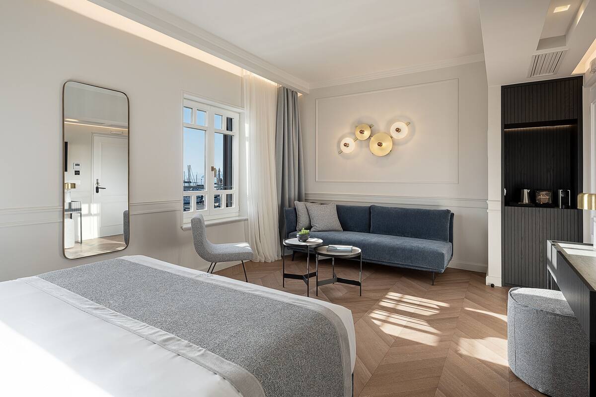 A modern hotel room with a bed, a sofa, a small table, a chair, a mirror, and a wall-mounted light fixture.