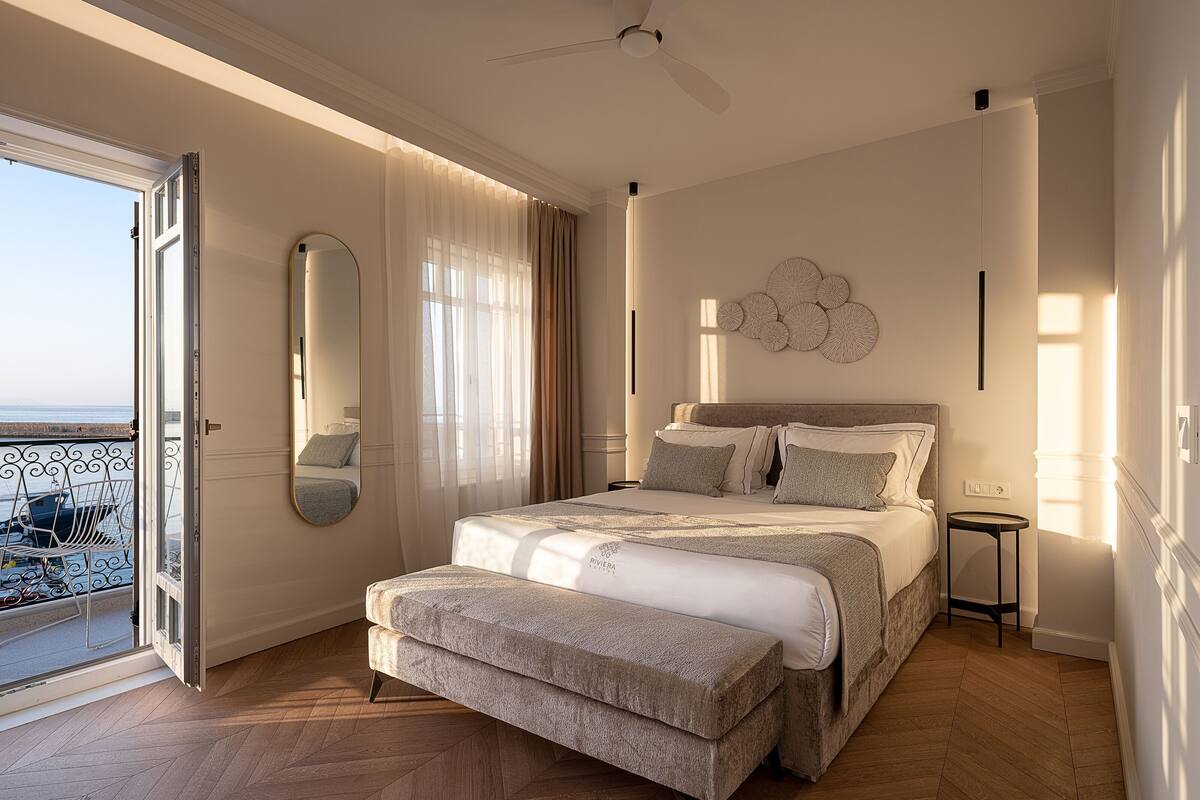 A bedroom with a bed, a bench, a ceiling fan, a round mirror, and a view of the outside.