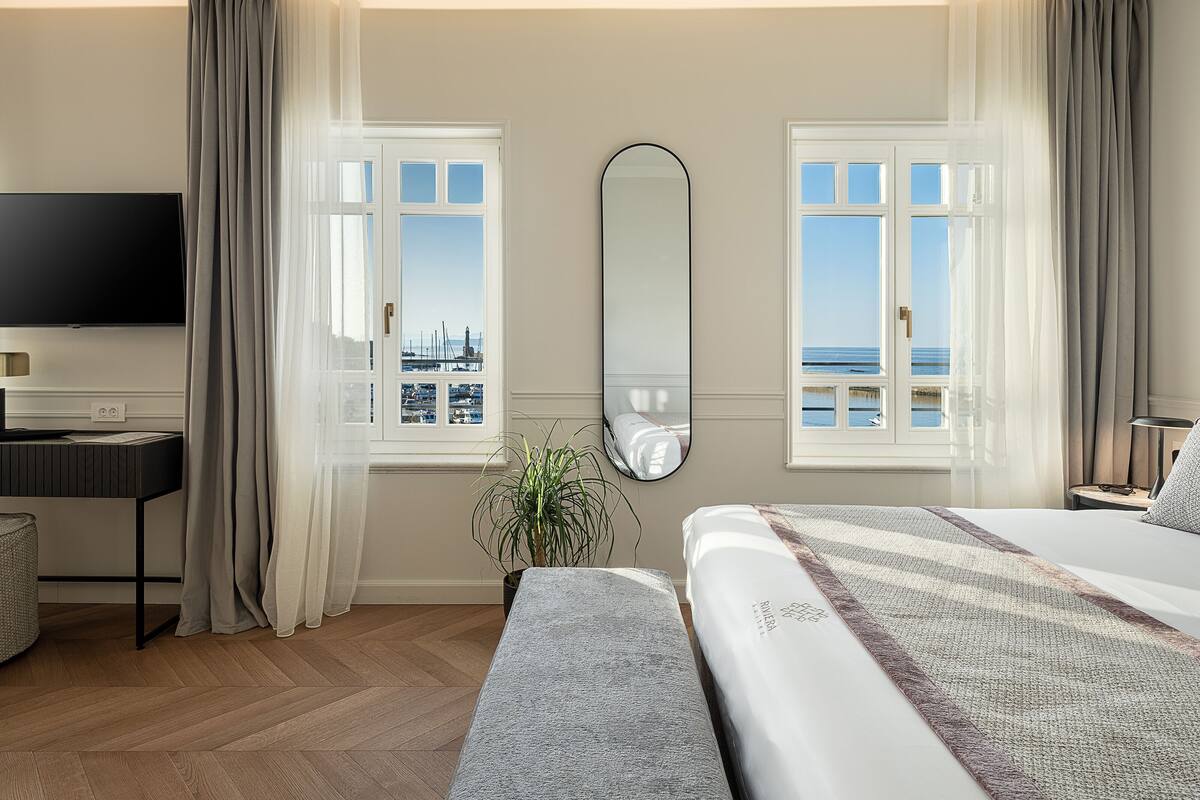 A hotel room with a bed, a TV, a large mirror, and a view of the sea.