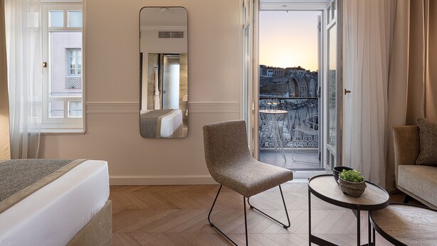 A hotel room with a bed, a chair, a small table with a plant, a mirror, and a view of the city through an open door. - Riviera Suites (Chania)