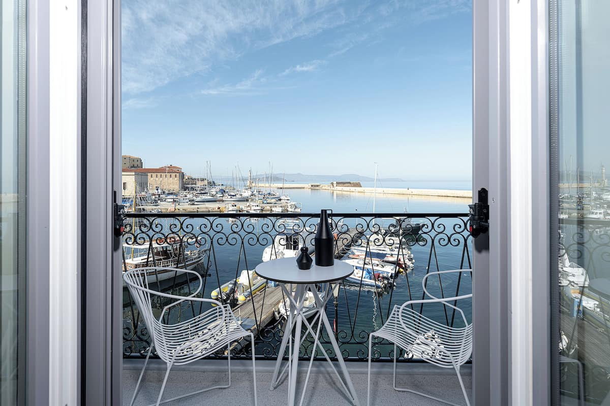 A balcony with two white chairs and a small table, overlooking a marina with boats and a distant building.