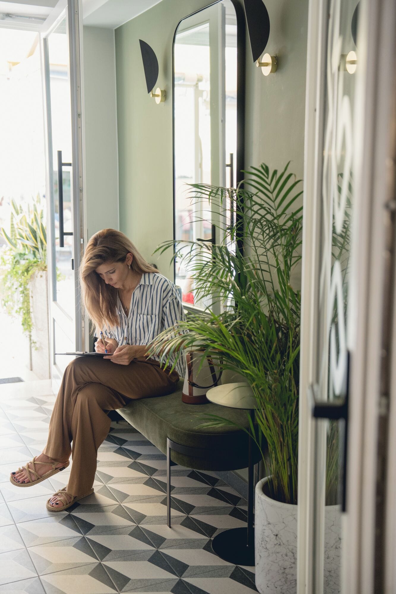 Una persona sentada en un banco en un espacio interior bien iluminado, con suelo de baldosas, una planta en maceta y un espejo en la pared.