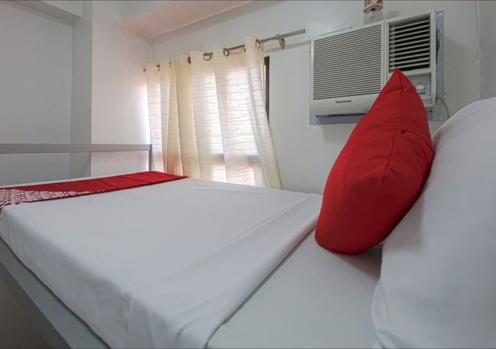 A hotel room with a bed, a red pillow, a white bedspread, and a window with curtains.