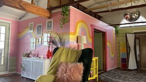 Une pièce avec un plafond haut, des poutres apparentes, un mur rose orné de motifs arc-en-ciel, un fauteuil vert, un coussin moelleux, une petite table avec un vase et une porte donnant sur une autre pièce.