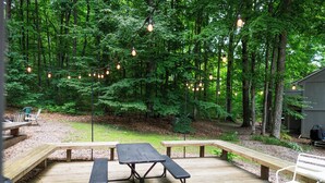 A wooden deck with picnic tables and benches, surrounded by a forested area. - The Ope! North Hideaway (Bellaire)