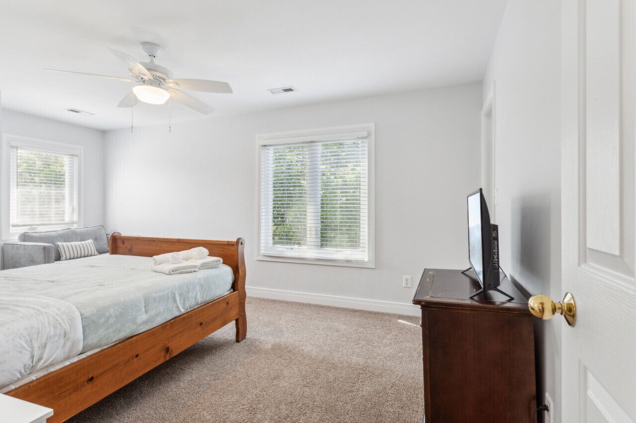 A bedroom with a wooden bed, a ceiling fan, a TV on a wooden dresser, and two windows with blinds.