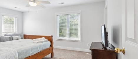 A bedroom with a wooden bed, a ceiling fan, a TV on a wooden dresser, and two windows with blinds.
