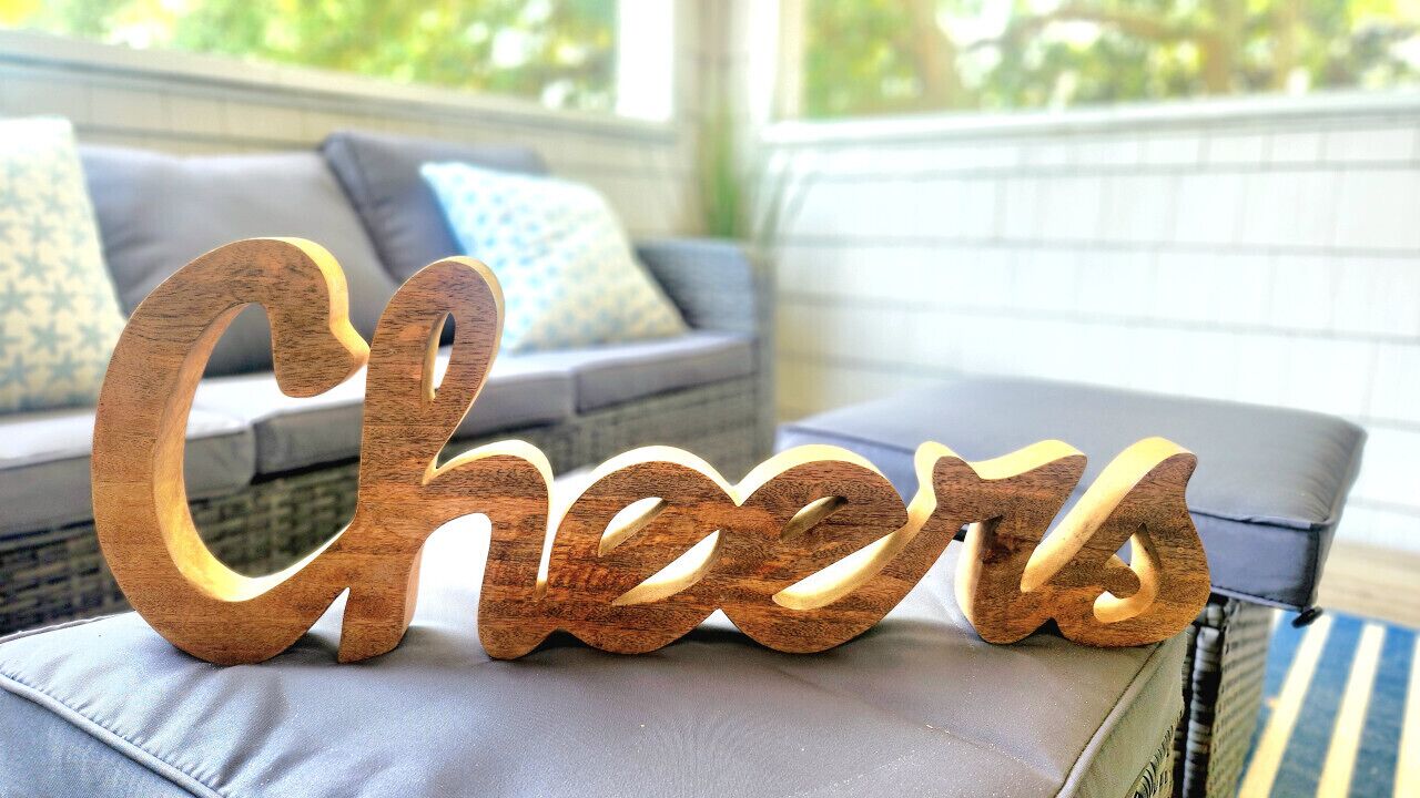 Un letrero de madera que dice 'Cheers' sobre una superficie acolchada en una habitación con un sofá gris y cojines estampados.