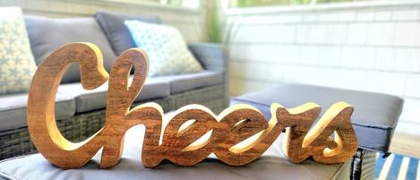 Un letrero de madera que dice 'Cheers' sobre una superficie acolchada en una habitación con un sofá gris y cojines estampados.