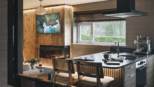 A modern kitchen with a wooden accent wall, a large TV, a fireplace, and a window with a view of greenery. - Scar View - Getaway for Two | Ridgestone (Kendal)