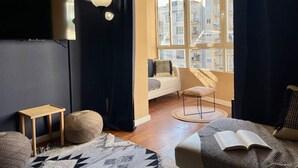 Una sala moderna con un sofá, una mesita y un sillón. Se ve un ventanal con vistas a edificios.