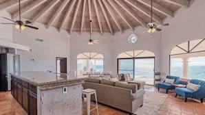 A spacious living area with a large island, a sofa, and a coffee table. There are two ceiling fans, a view of the ocean through large windows, and a kitchen with a refrigerator.