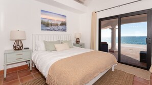A bedroom with a bed, nightstands, a painting, and a view of the sea through a glass door.