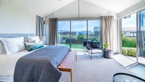 A modern bedroom with a large bed, a bench, a chair, and a view of the outdoors. - Serenity on Ruby Ridge (Wānaka)