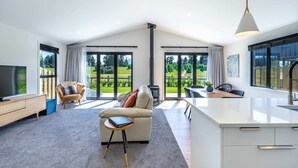 A spacious living area with a sofa, a coffee table, a TV, and a fireplace. There are large windows offering a view of a garden.