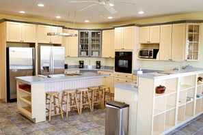 Une cuisine avec des meubles de couleur bois clair, des appareils en acier inoxydable, un îlot central doté de tabourets et un ventilateur de plafond.
