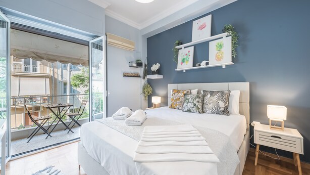 A modern bedroom with a large bed, a bedside table with a lamp, and a balcony with outdoor furniture. - City Nest Apartment by Cloudkeys (Athens)