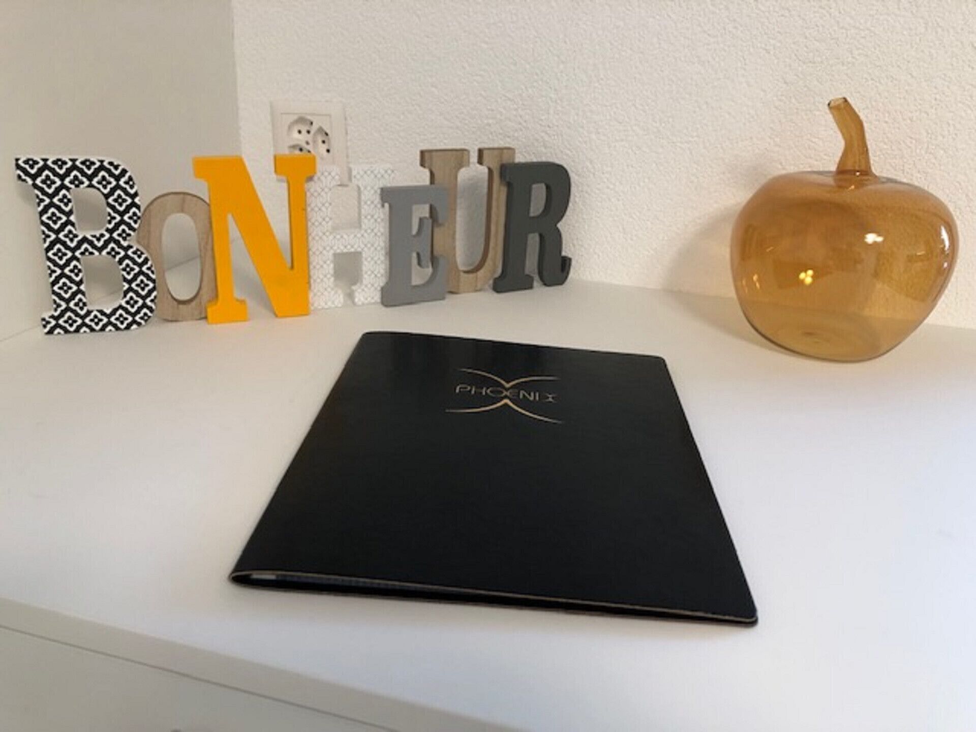 A black folder with 'PHOENIX' logo on a white surface, surrounded by decorative letters and a pumpkin.
