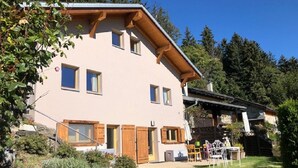 A two-story building with a sloped roof, wooden shutters, and a small patio area with outdoor furniture. - My Cosy Flat in the Mountains (Anzère)