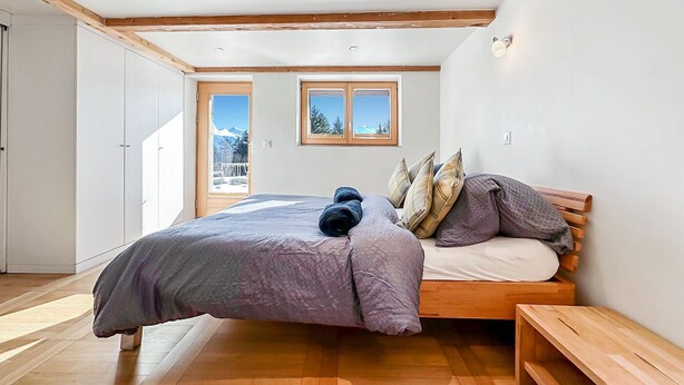 A bedroom with a bed, wooden headboard, two windows, and a balcony with a view of mountains. - My Cosy Flat in the Mountains (Anzère)