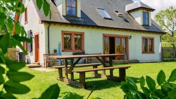 A two-story house with a sloped roof, a visible chimney, and a front porch with a bench. - Country farmhouse sleeps 9 with enclosed garden and games barn. (Blairgowrie, Perthshire)