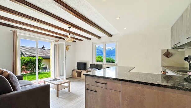 A modern kitchen with a large island, a dining area with a sofa and table, and a view of mountains through the windows. - Chez Tonton à 5mns de Sion (Sion)