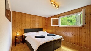 A bedroom with a wooden paneled wall, a large bed with a dark headboard, a bedside table with a lamp, and a window with a view of greenery. - Chez Tonton à 5mns de Sion (Sion)
