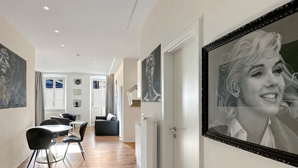 Un couloir au sol en bois, un coin repas avec une table et des chaises, et un portrait encadré accroché au mur.
