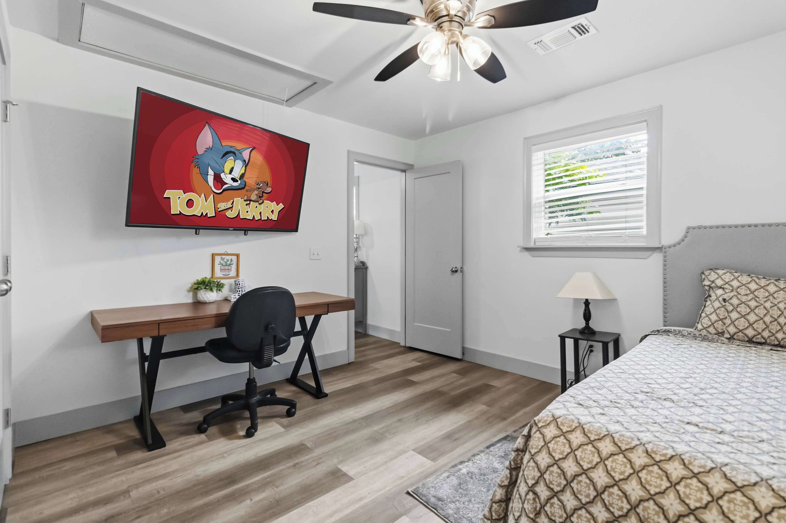 A bedroom with a bed, a desk with a chair, a ceiling fan, a window with blinds, and a wall-mounted TV displaying 'Tom and Jerry'.