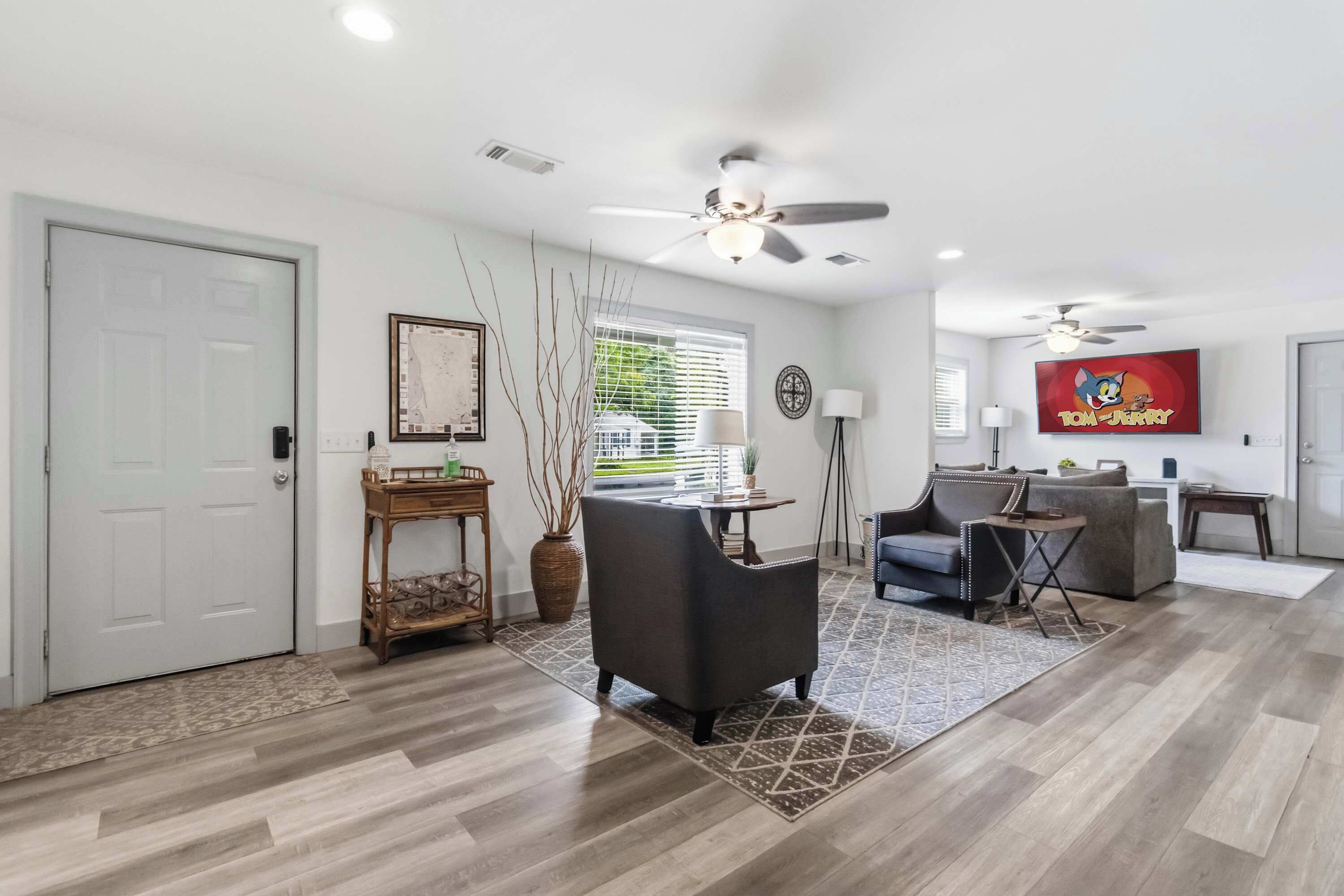 A spacious living room with a ceiling fan, a television displaying 'Tom and Jerry', and a large window with blinds.