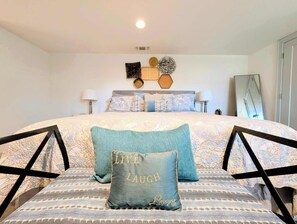 Un dormitorio con una cama grande, varios almohadones, una mesita de noche, una lámpara y un espejo colgado en la pared.