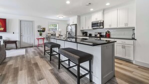 A modern kitchen with white cabinetry, a central island with bar stools, and a dining area with a red chair. - Spacious Renovated Home with Fenced Yard (Springdale)