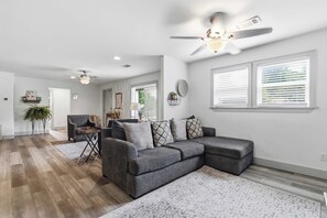 A modern living room with a gray sectional sofa, a ceiling fan, and a large window with blinds. - Spacious Renovated Home with Fenced Yard (Springdale)